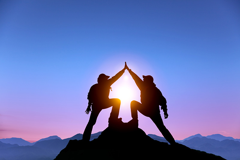 Two people high fiving at the top of peak with the sun behind them